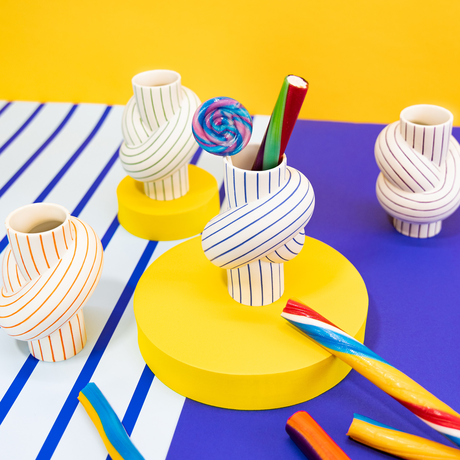 I quattro vasi Node Stripes, in parte riempiti di lecca-lecca e bastoncini di zucchero, sono affiancati da uno sfondo astratto in giallo, bianco e viola.